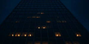 Glass office tower facade at dusk with scattered lit windows, viewed from street level